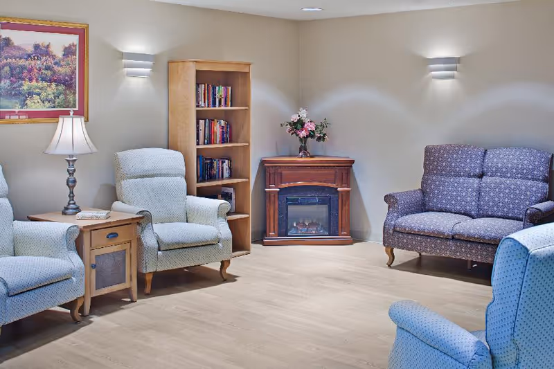 A cozy living room area with four upholstered armchairs and a loveseat arranged around a small wooden electric fireplace with a flower vase on top. There is a wooden bookshelf filled with books, a side table with a lamp and a book, and a framed landscape painting on the wall. The room has light-colored walls and wood flooring.