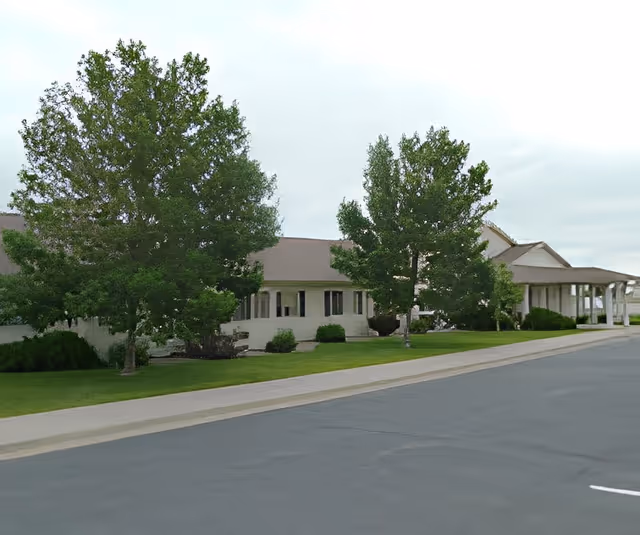 Exterior view of a single-story building with a covered entrance, surrounded by green trees and well-maintained grass, under an overcast sky.