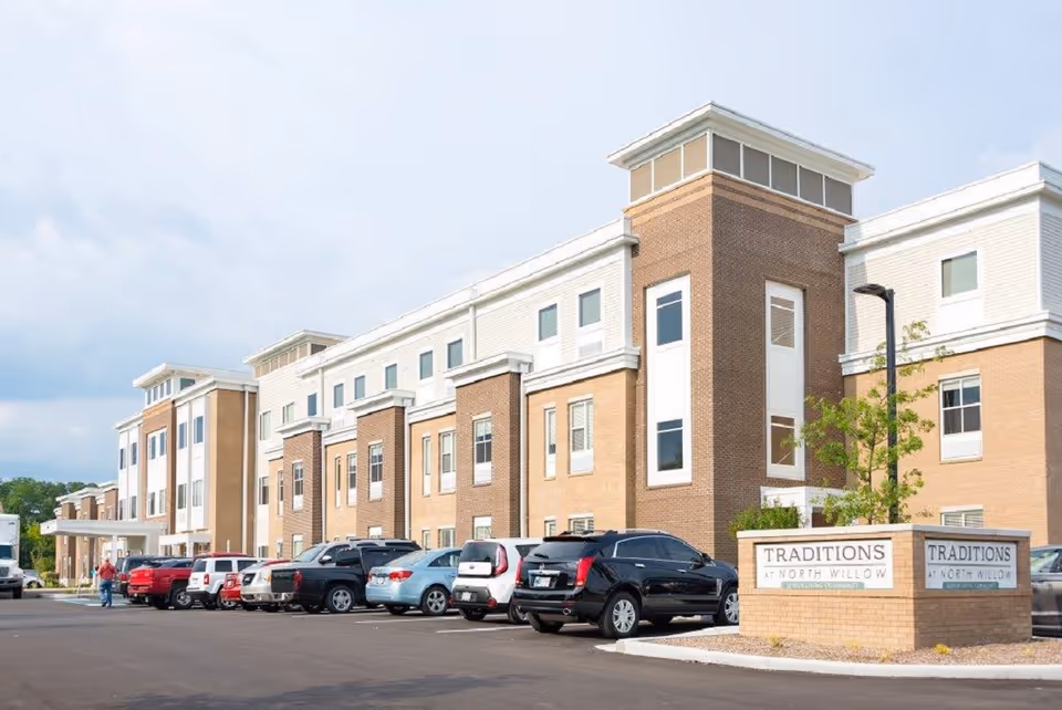 Exterior view of a three-story senior living facility named Traditions at North Willow with a parking lot in front containing several parked cars and a person walking near the entrance.