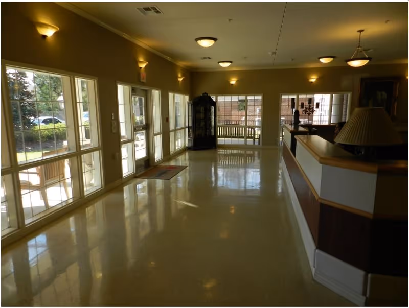 A spacious, well-lit interior hallway or lobby area with large windows on the left side allowing natural light to enter. There is a wooden reception desk on the right with a lamp on it, and a glass display cabinet at the far end near a bench. The floor is shiny and reflective, and wall-mounted lights and ceiling fixtures provide additional lighting.