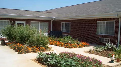 Outdoor courtyard area of a brick building with multiple windows and doors, featuring flower beds with various colorful flowers and plants along a paved walkway.