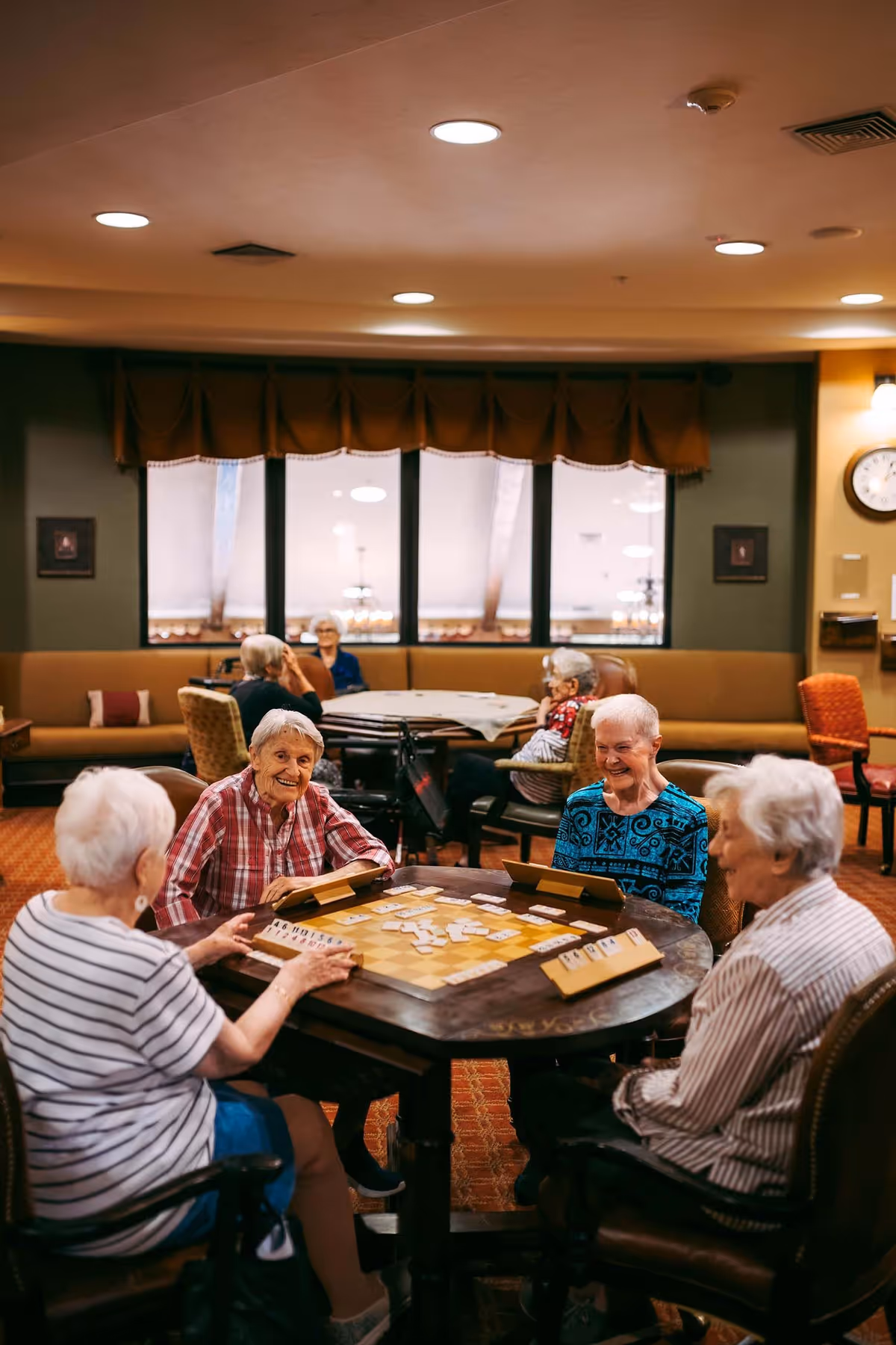 A group of elderly women sitting around a round table playing a board game in a cozy, well-lit common room with large windows and comfortable seating. Other elderly individuals are seated at tables in the background, engaging in conversation or activities.
