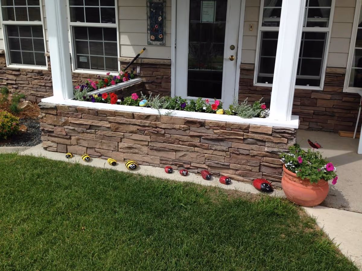 Front porch area of a building with stone facade and white columns. There are colorful flowers planted in a raised flower bed and a large terracotta pot with flowers on the right side. Decorative painted rocks resembling bees and ladybugs are lined up along the edge of the concrete walkway in front of the flower bed. The porch has windows and a glass door.