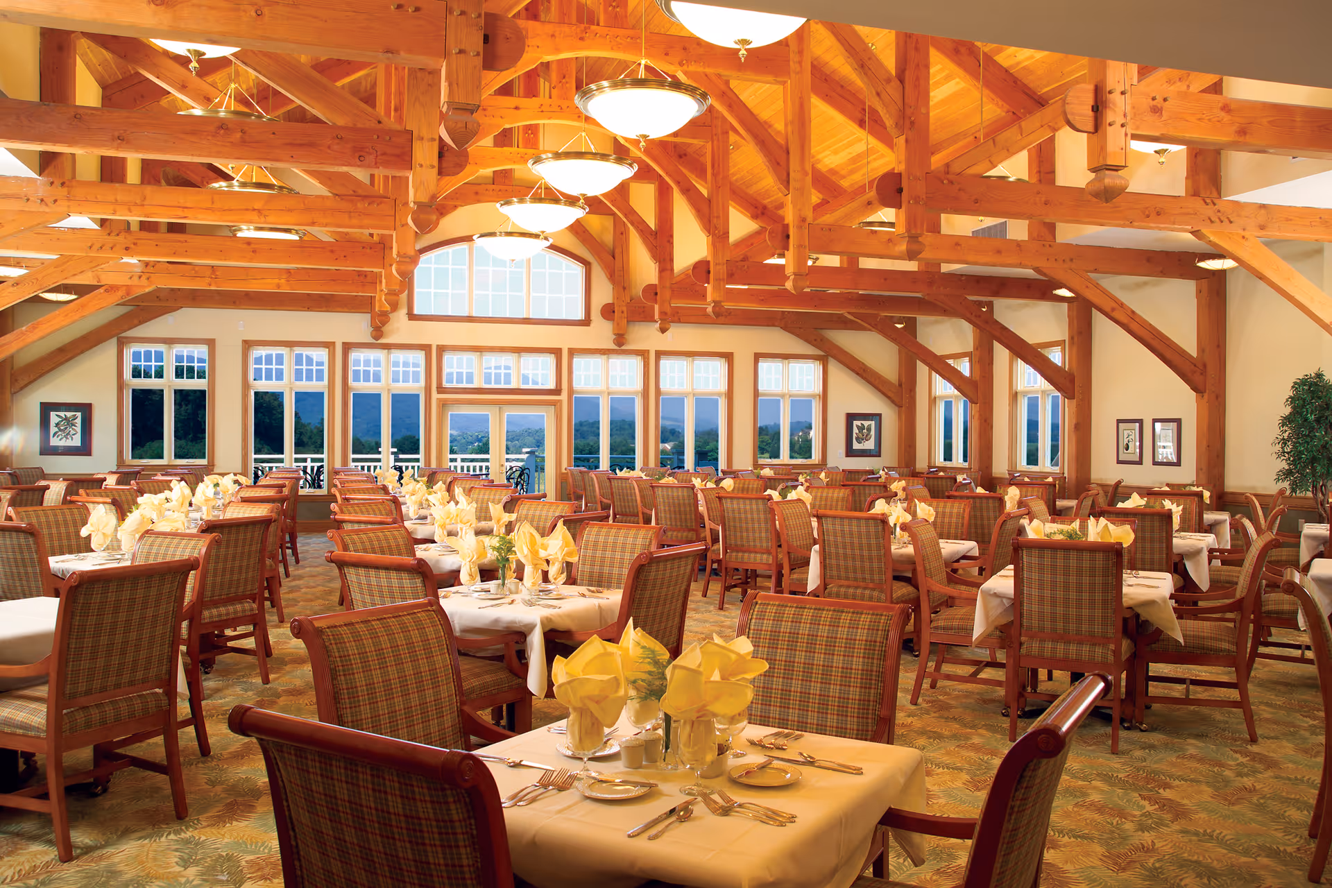 Spacious dining room with multiple tables set with white tablecloths, yellow folded napkins, and silverware. The room features large windows with a scenic view outside, wooden beams on the ceiling, and warm lighting fixtures.