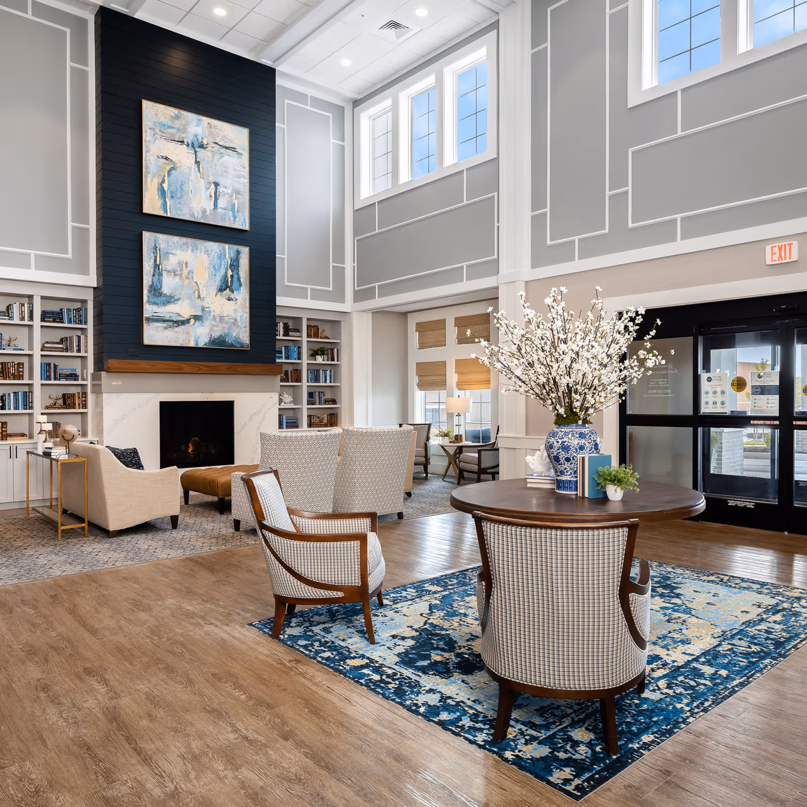 Bright senior living lobby with seating areas, a fireplace flanked by bookshelves, and a round table with a vase of flowers.