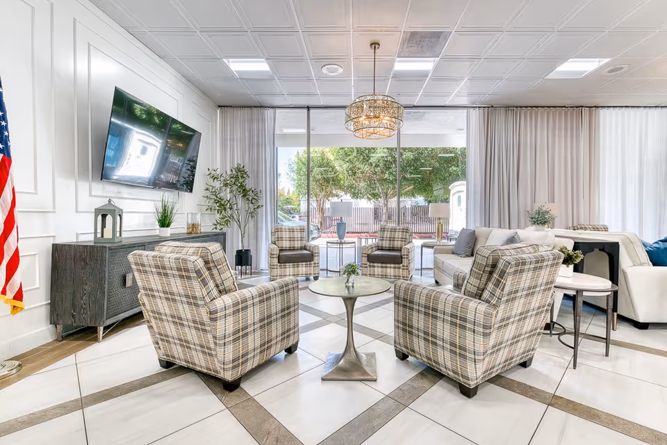 A bright and modern living room area with four plaid armchairs arranged around a small round table. There is a large flat-screen TV mounted on the wall above a dark wooden cabinet, an American flag to the left, and large windows with sheer curtains letting in natural light. Additional seating includes a white sofa with cushions and side tables with plants.