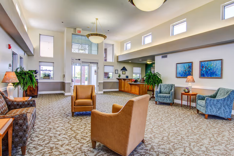 A spacious senior living community lobby with patterned carpet, several armchairs and sofas arranged for seating, potted plants, two framed paintings on the wall, and a reception desk near the entrance with large windows and glass doors allowing natural light.