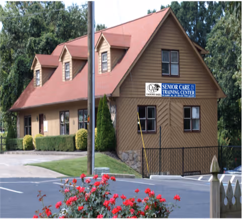 Exterior view of a two-story brown building with a red roof, surrounded by greenery and a parking lot. The building has a sign that reads 'Cherokee Angel Senior Care & Training Center'. There are red flowers in the foreground near a fence.