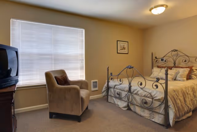 A cozy bedroom with a decorative metal bed frame, a neatly made bed with multiple pillows, a beige armchair with a dark cushion, a window with closed blinds, a framed picture on the wall, and a small heater mounted on the wall.