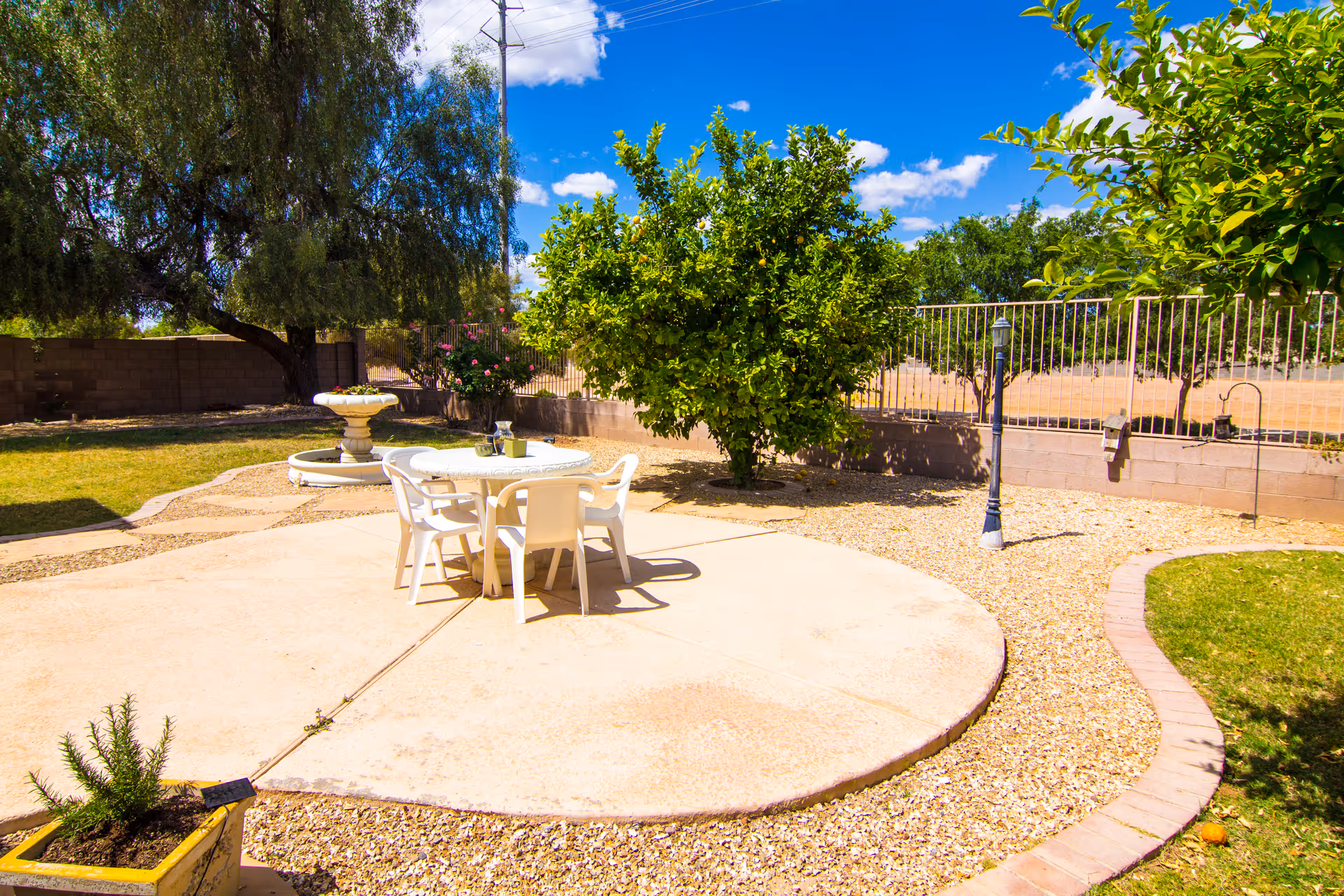 Outdoor patio area with a round concrete platform holding a white table and four white chairs. Surrounding the patio are gravel and green grass areas, with trees and a birdbath in the background under a blue sky with some clouds.