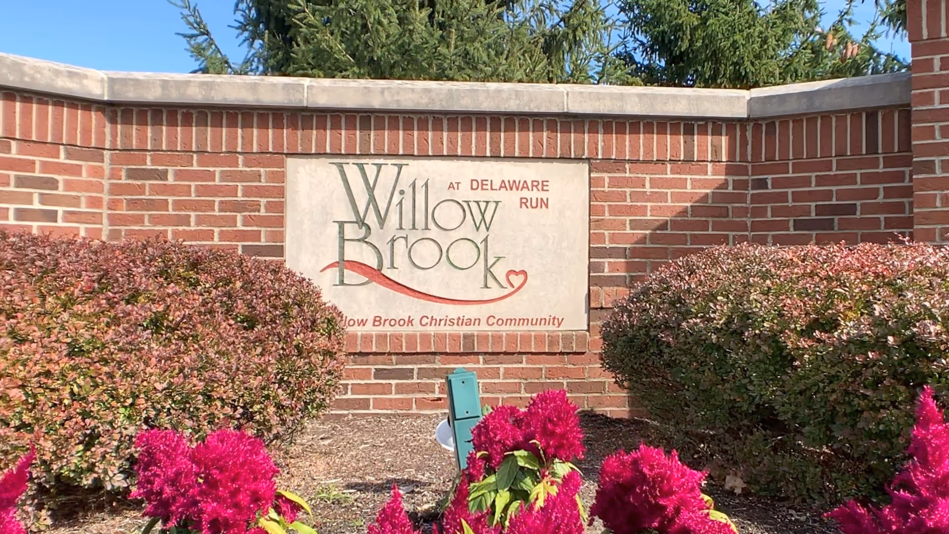 Brick entrance sign that reads 'Willow Brook at Delaware Run' set in landscaping with shrubs and pink flowers.