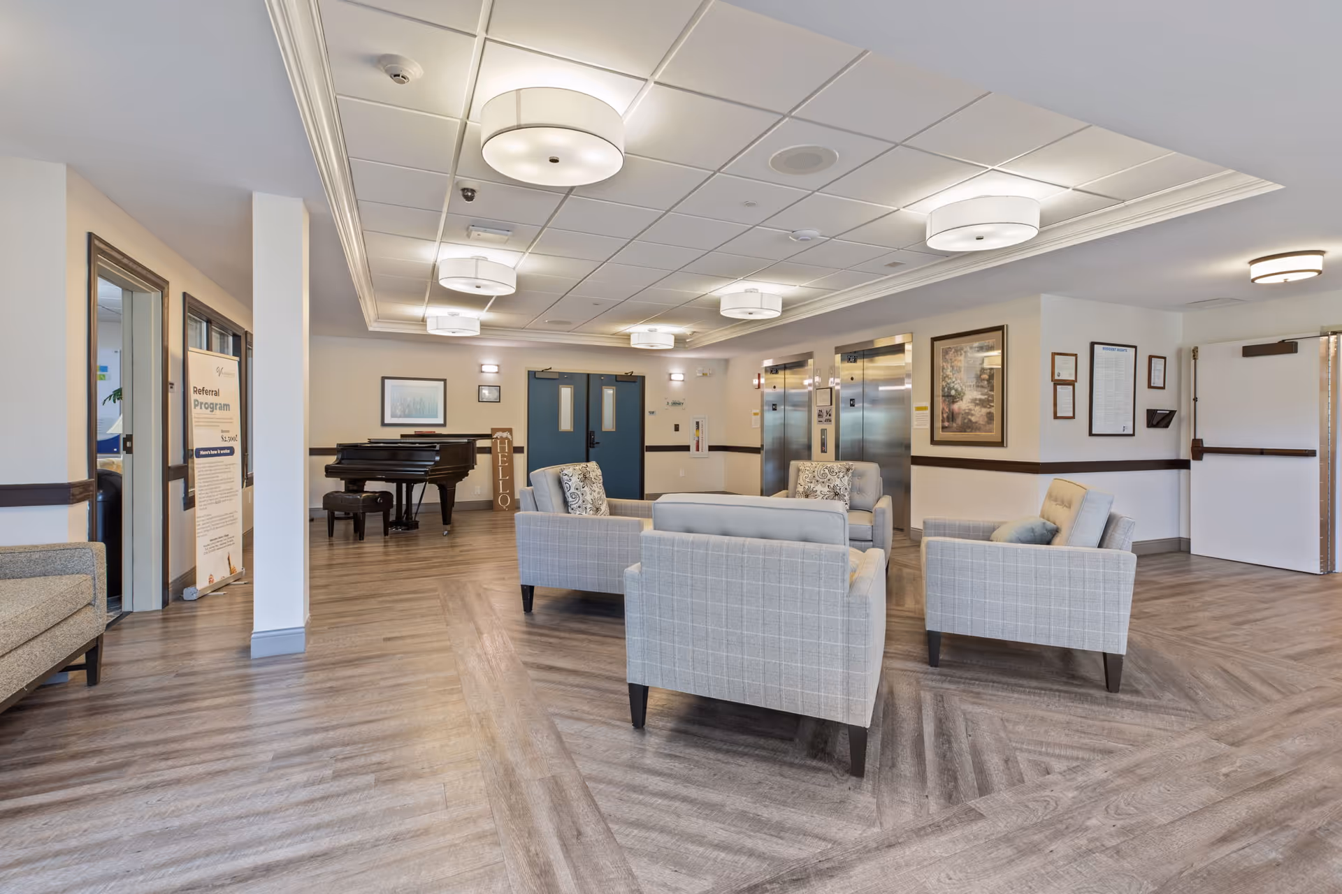 Open senior living lounge with upholstered chairs arranged around a seating area, a piano, and elevators in the background.