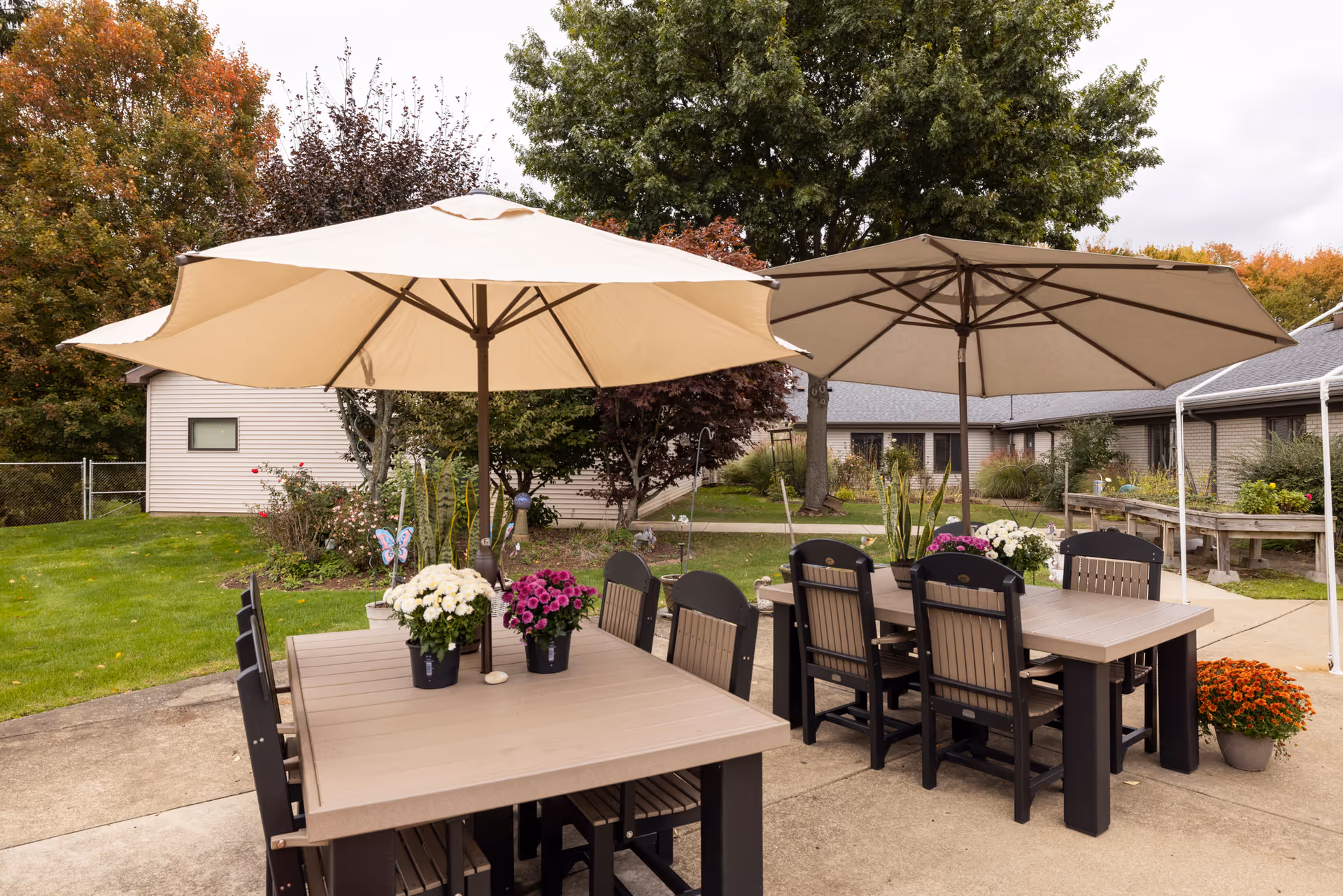 Outdoor patio area at a senior care facility with two large beige umbrellas shading two tables surrounded by chairs. Each table has potted flowers on top. The patio is surrounded by green grass, trees with autumn foliage, and single-story buildings in the background.