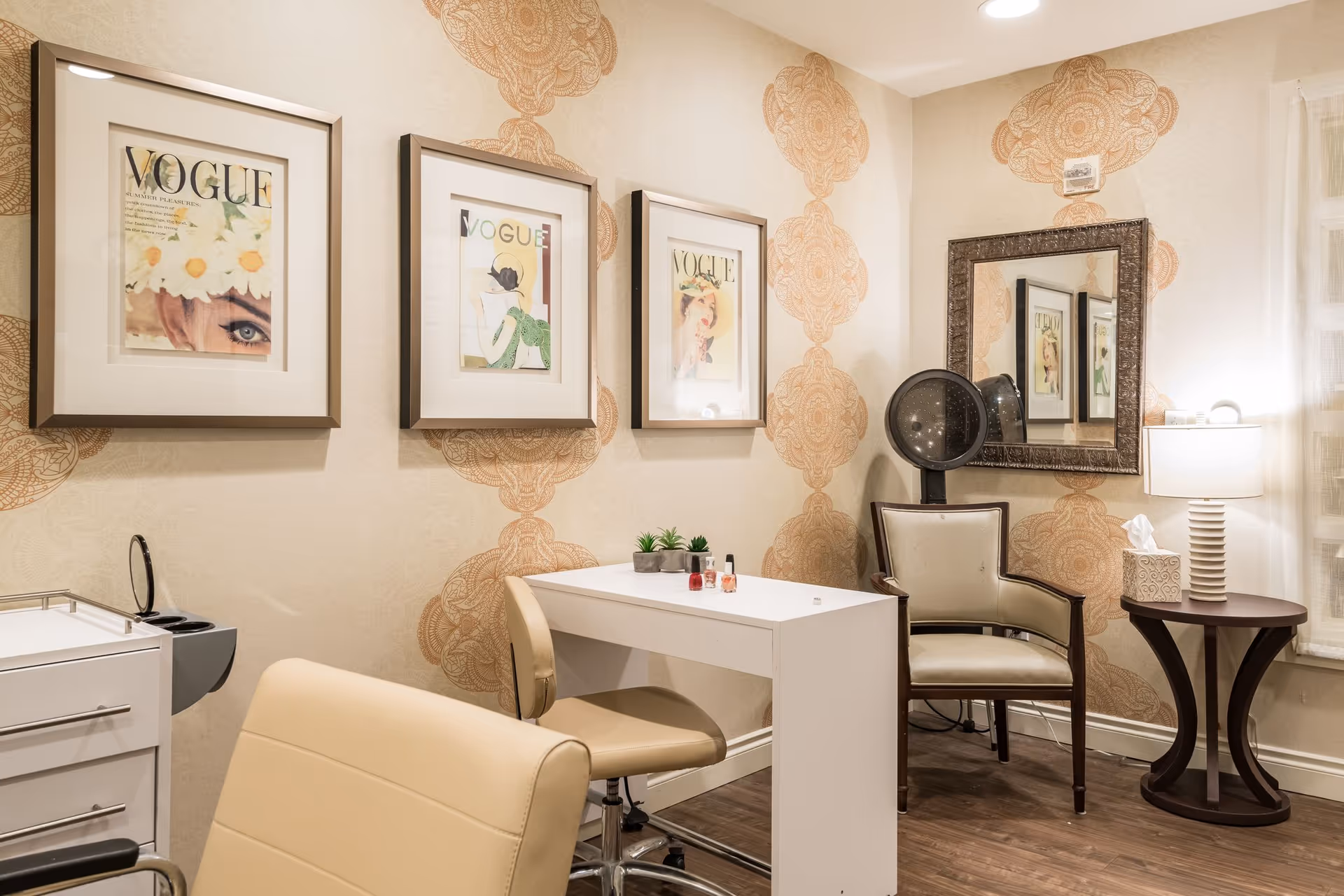 A cozy salon-style interior with a manicure table, chairs, framed Vogue prints, a wall mirror and a hooded hair dryer.