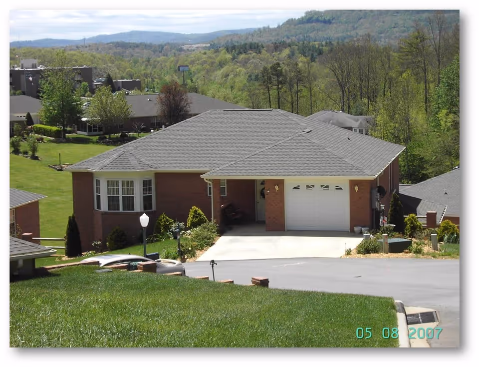 A single-story brick building with a gray shingled roof, a white garage door, and a small covered porch area. The building is surrounded by green grass, shrubs, and trees, with a paved driveway leading up to the garage. In the background, there are more buildings and a wooded hillside under a partly cloudy sky.