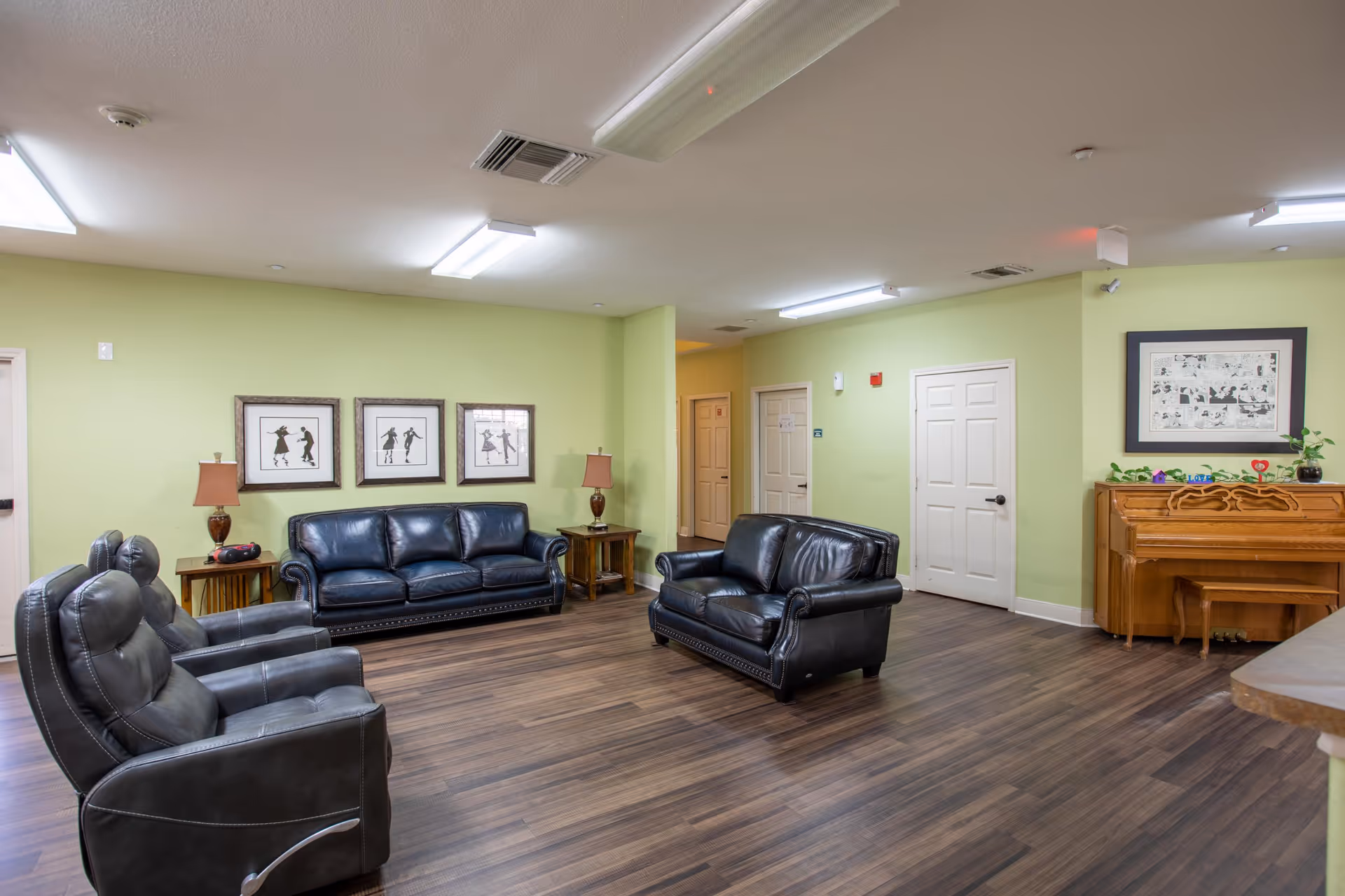 A spacious living room with light green walls and wood flooring. The room features two black leather sofas and two black leather recliners arranged around the space. There are two wooden side tables with lamps on either side of one sofa. On the right side of the room, there is a wooden piano with a bench and some decorative items on top. Three framed silhouette art pieces hang on the wall above one sofa, and another framed artwork is above the piano. Several white doors are visible in the background under fluorescent ceiling lights.