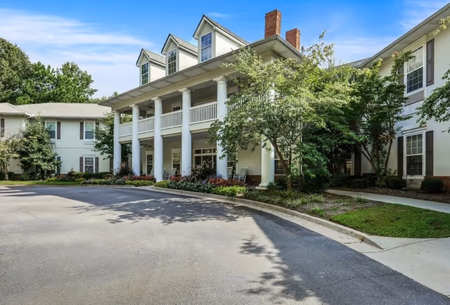 Two-story senior living building with white columns, a covered front porch and a circular driveway surrounded by trees and landscaping.
