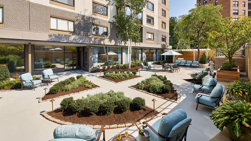 Sunlit courtyard patio at a senior living facility with cushioned chairs, planted garden beds, and umbrella-shaded tables outside a multi-story building.