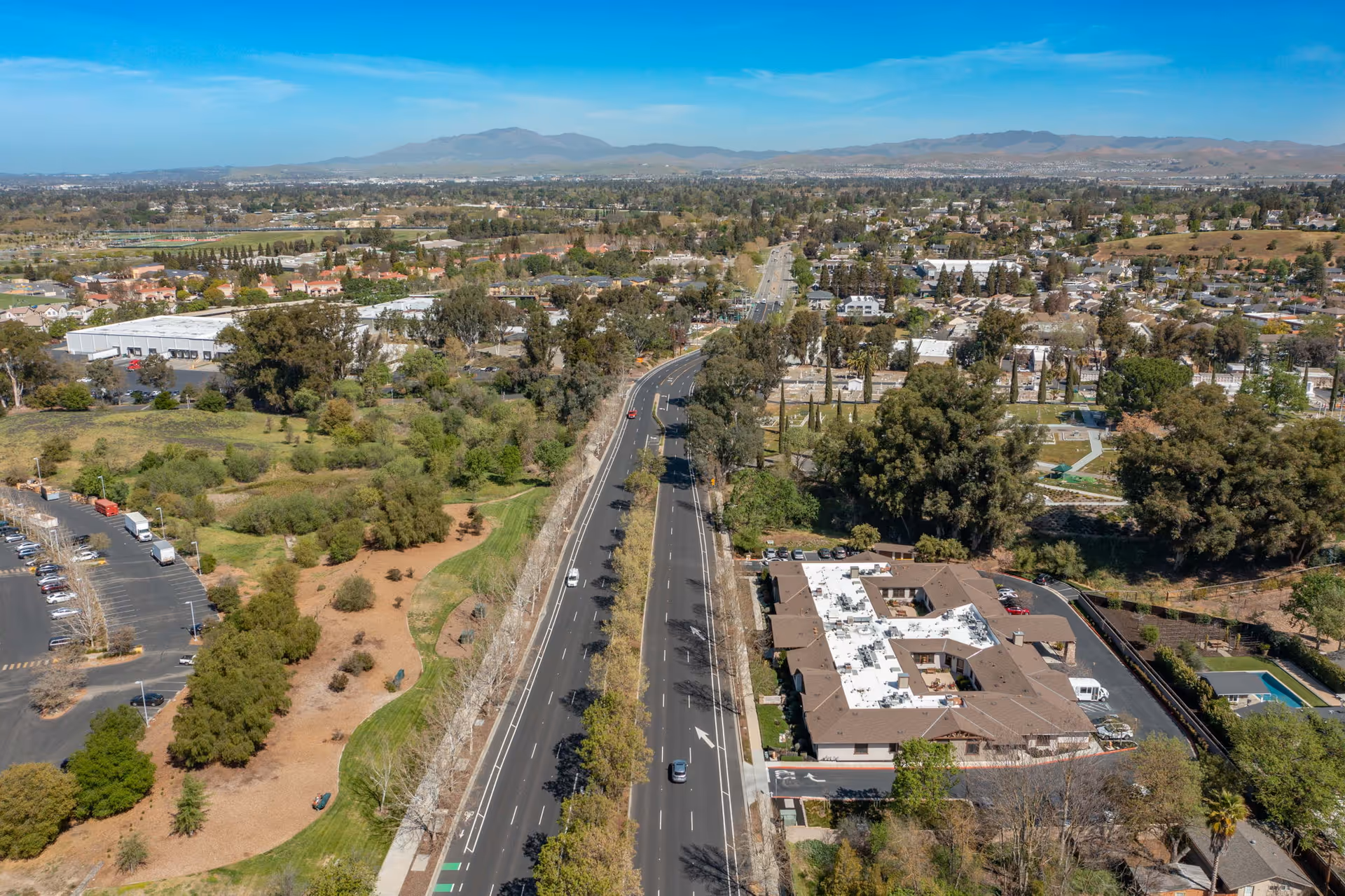 Aerial view of a suburban area featuring a multi-lane road running through the center, surrounded by trees, green spaces, and residential and commercial buildings. A large building complex with a white roof is visible on the right side of the road, likely the Sunol Creek Memory Care facility. Mountains are visible in the distant background under a clear blue sky.