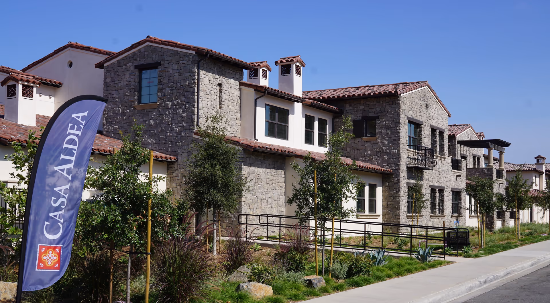 Exterior view of a senior living facility building with stone and stucco walls, red tile roof, multiple windows, and small balconies. There is a sidewalk and landscaped greenery in front, along with a blue flag banner that reads 'CASA ALDEA'.