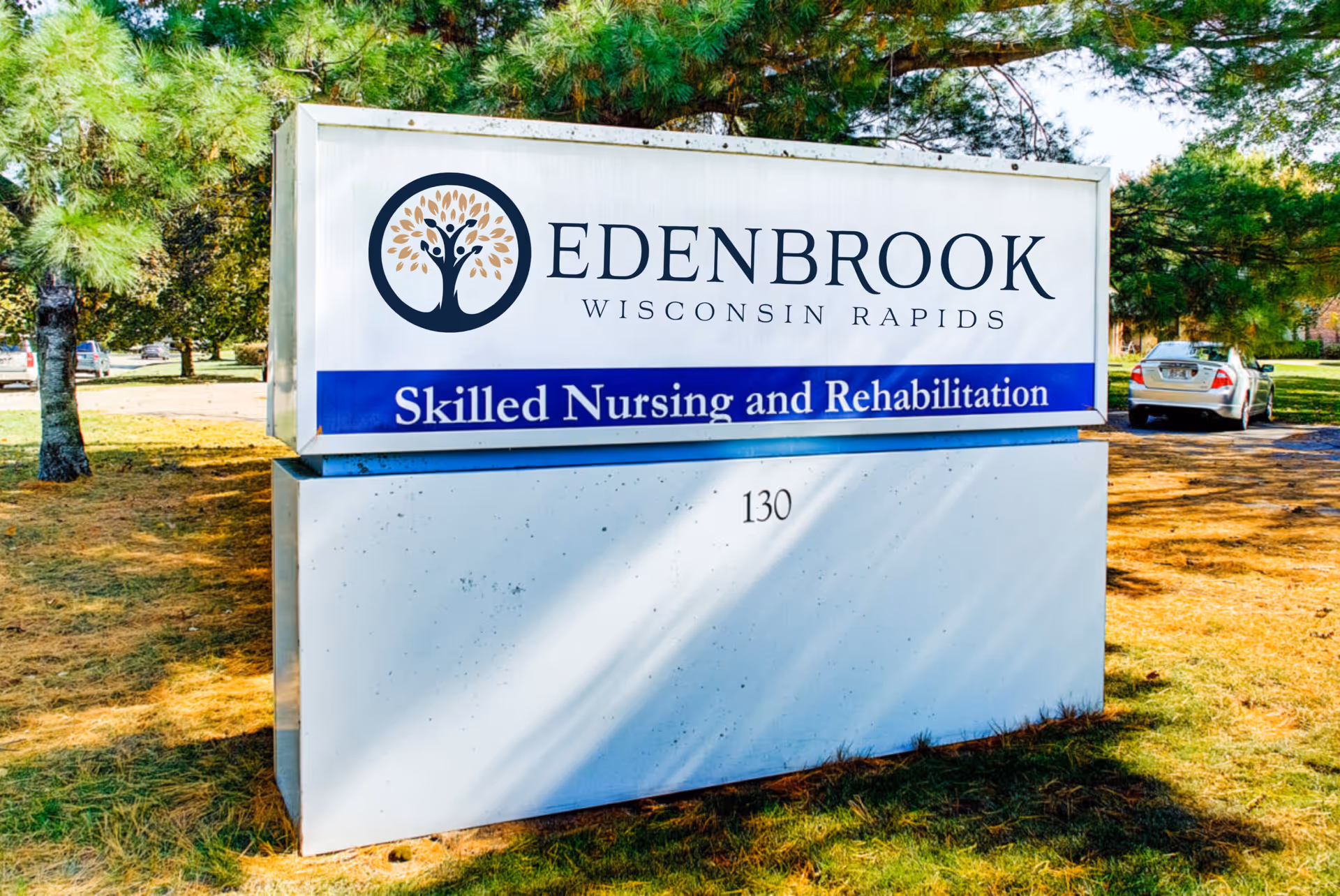 Outdoor sign for Edenbrook Wisconsin Rapids Skilled Nursing and Rehabilitation facility, surrounded by trees and parked cars in the background.