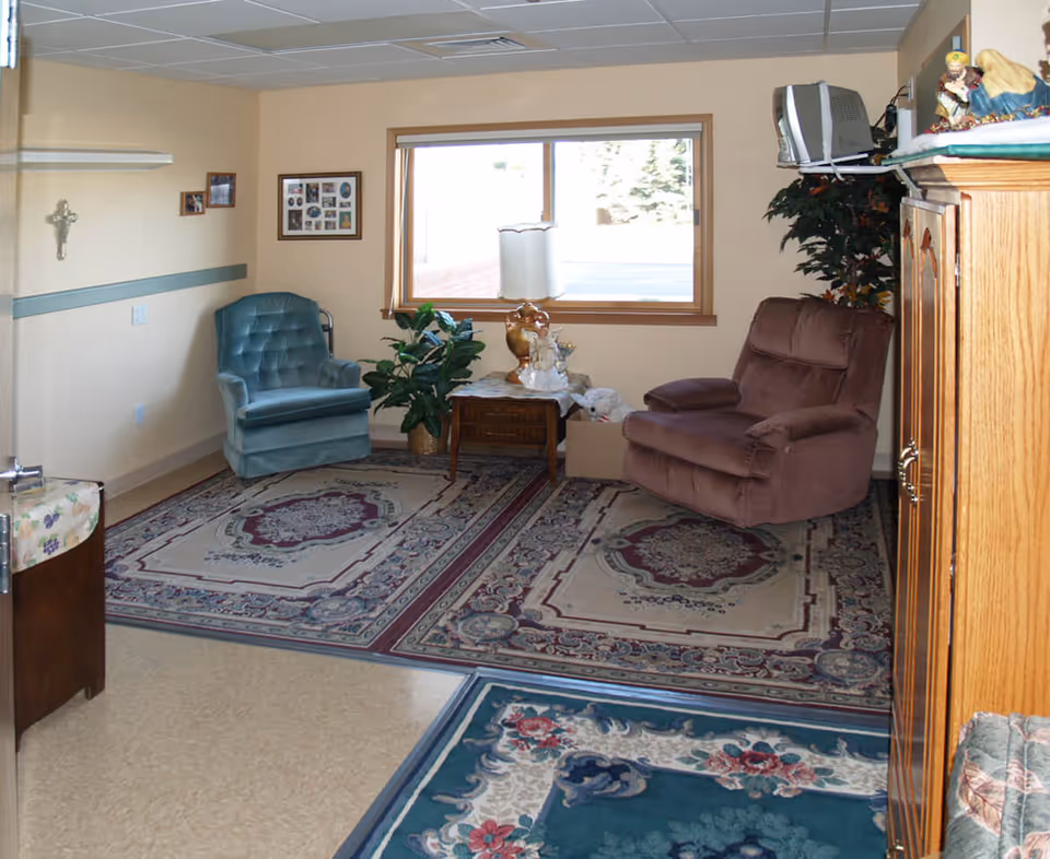 A cozy sitting area in a senior living facility with a blue armchair and a brown recliner on patterned area rugs. There is a wooden side table with a lamp and decorative items between the chairs, a window letting in natural light, framed pictures on the wall, a crucifix, and a wooden cabinet with a small TV on top. A potted plant is placed near the window.