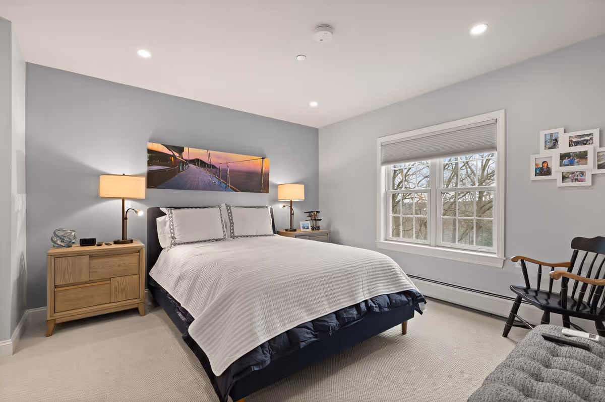 A neatly arranged bedroom with a large bed covered in white bedding and two pillows. There are two wooden nightstands on either side of the bed, each with a lamp. Above the bed is a panoramic wall art depicting a sunset over a dock. A window with white blinds lets in natural light, and a black wooden chair and a gray cushioned bench are placed near the window. Family photos are displayed on the wall next to the window.