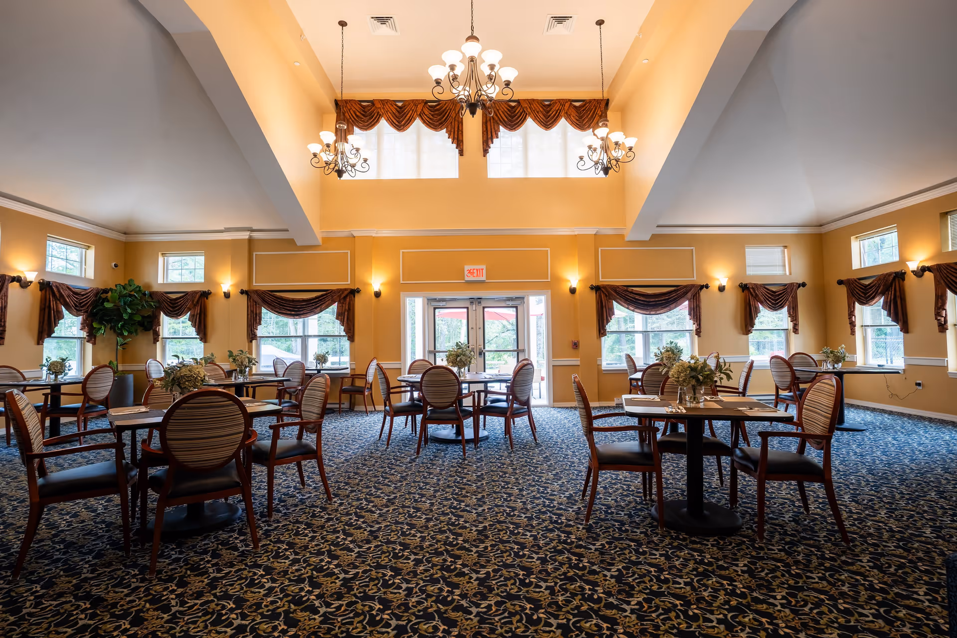 A spacious dining room with multiple round tables and chairs arranged neatly. The room has high ceilings with three ornate chandeliers hanging down. Large windows with brown drapes allow natural light to fill the room. The carpet has a dark blue and gold intricate pattern, and there are floral centerpieces on each table. Double glass doors lead outside, and the walls are painted a warm yellow color.