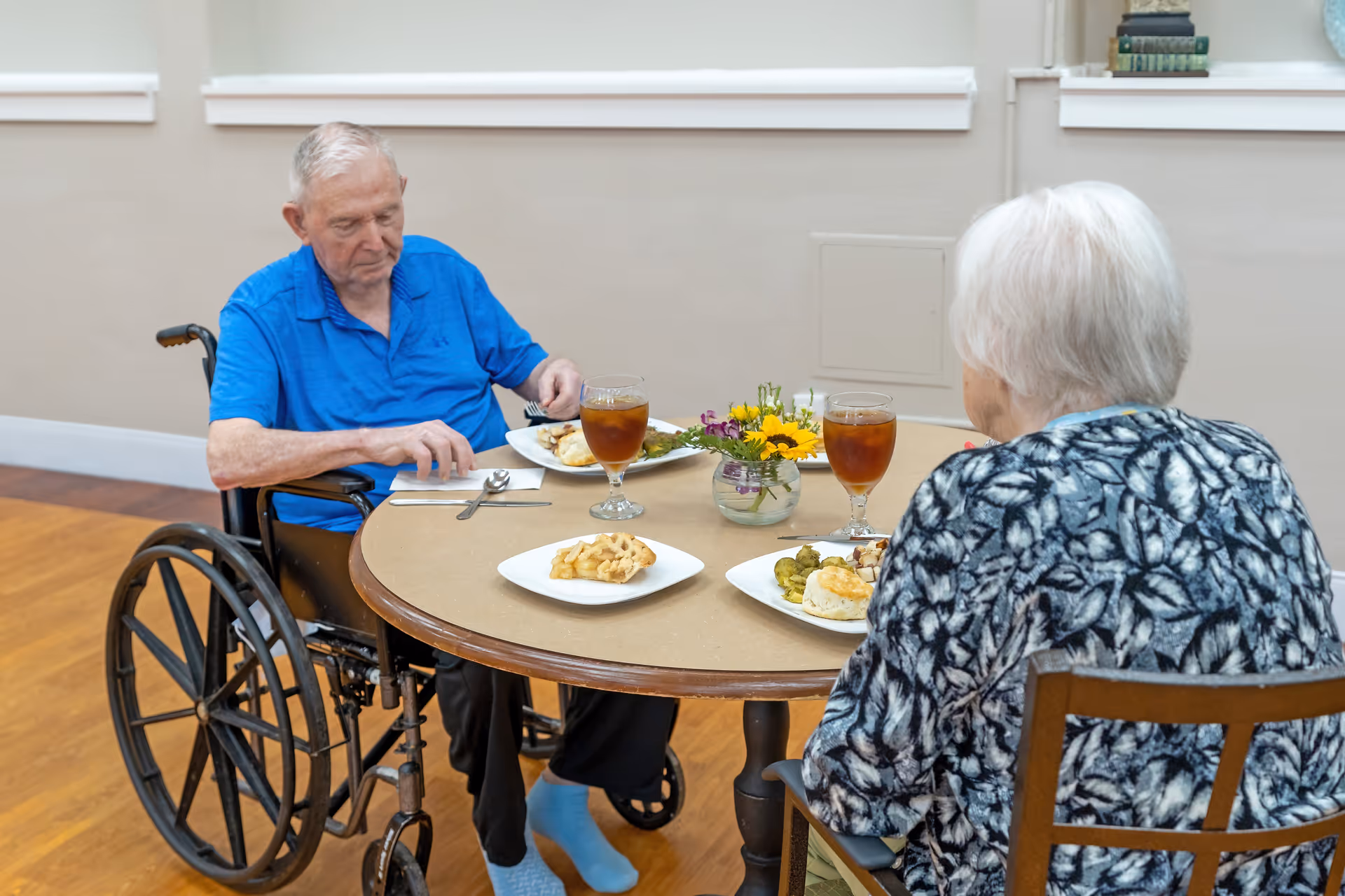 Two elderly individuals sitting at a round table having a meal together. One person is in a wheelchair wearing a blue shirt, and the other has white hair and is wearing a patterned top. The table has plates of food, glasses of iced tea, and a small vase with flowers. The setting appears to be indoors with wooden flooring and light-colored walls.