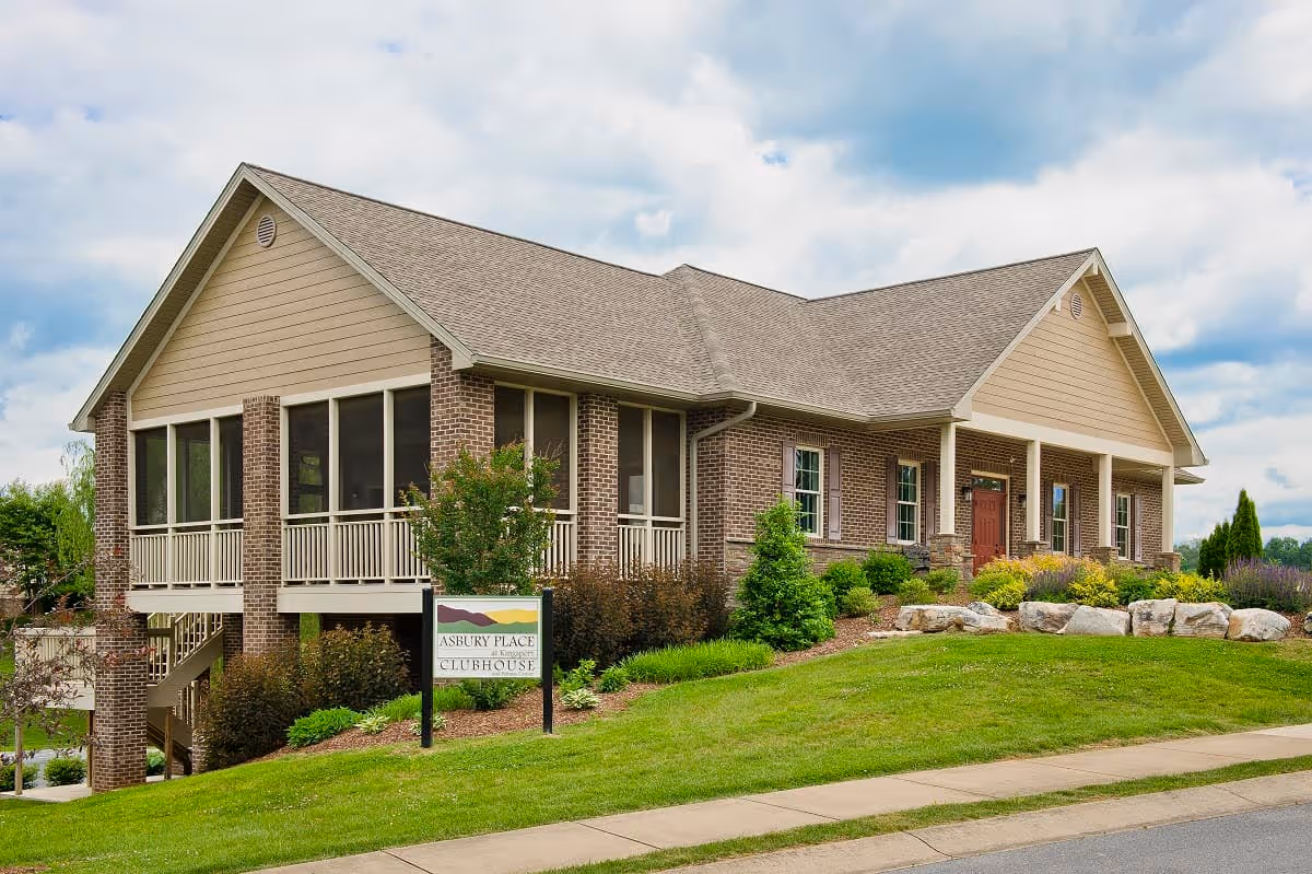 Exterior view of a single-story brick and siding building with a gabled roof, surrounded by a well-maintained lawn and landscaping. The building has a porch with railings and a sign in front that reads 'Asbury Place Clubhouse'. The sky is partly cloudy.