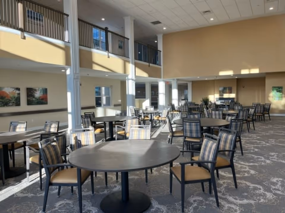 A spacious dining room with multiple round and rectangular tables surrounded by chairs with striped upholstery. The room has high ceilings, large windows allowing natural light to fill the space, and a second-floor balcony overlooking the area. The walls are painted beige and decorated with framed artwork.