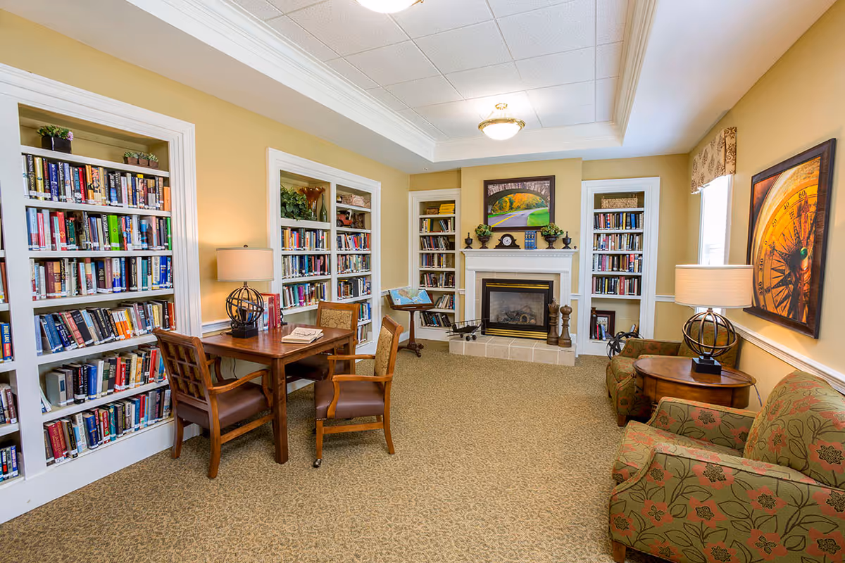 A cozy senior living facility library and sitting area with built-in bookshelves filled with books, a wooden table with chairs, two upholstered armchairs with floral patterns, two table lamps, a fireplace with a mantel decorated with plants and a clock, and framed artwork on the walls.