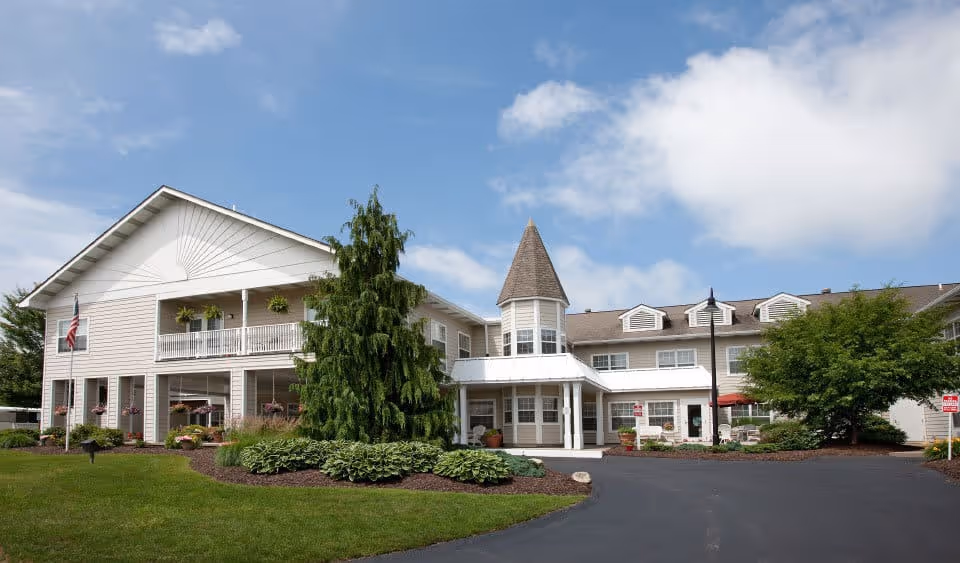 Exterior view of a senior living facility building with beige siding, a peaked turret, and a covered entrance. The building is surrounded by well-maintained landscaping including green grass, bushes, and trees. The sky is partly cloudy with blue patches visible.