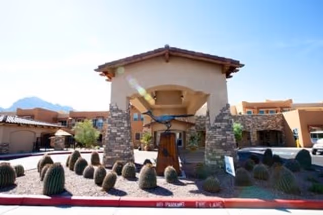 Exterior view of Desert Springs Gracious Retirement Living facility showing a covered entrance with stone pillars, desert landscaping with cacti, and a clear blue sky in the background.