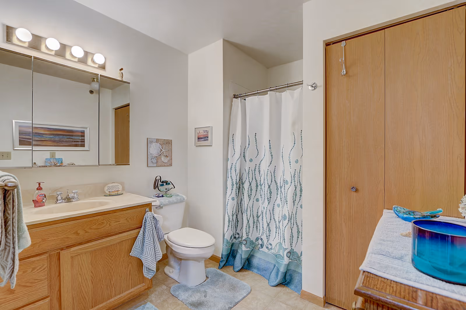 A clean bathroom with a wooden vanity featuring a sink and mirror above it. There is a toilet next to the vanity, and a shower with a white curtain decorated with blue and green patterns. The floor has light-colored tiles and there are blue bath mats. A wooden closet door is visible on the right side, and a small table with decorative items is partially visible in the foreground.
