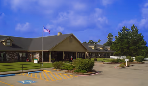 Exterior view of a single-story senior living facility building with a brick and siding facade, an American flag on a flagpole, landscaped bushes, and a parking lot with a handicapped parking space in front.