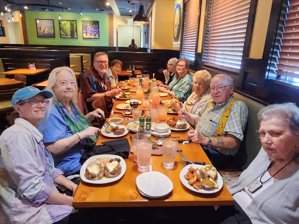 A group of elderly people sitting around a long wooden table in a restaurant, enjoying a meal together. The table is filled with plates of food, glasses of drinks, and condiments. The restaurant has wooden blinds on the windows and framed pictures on the walls.
