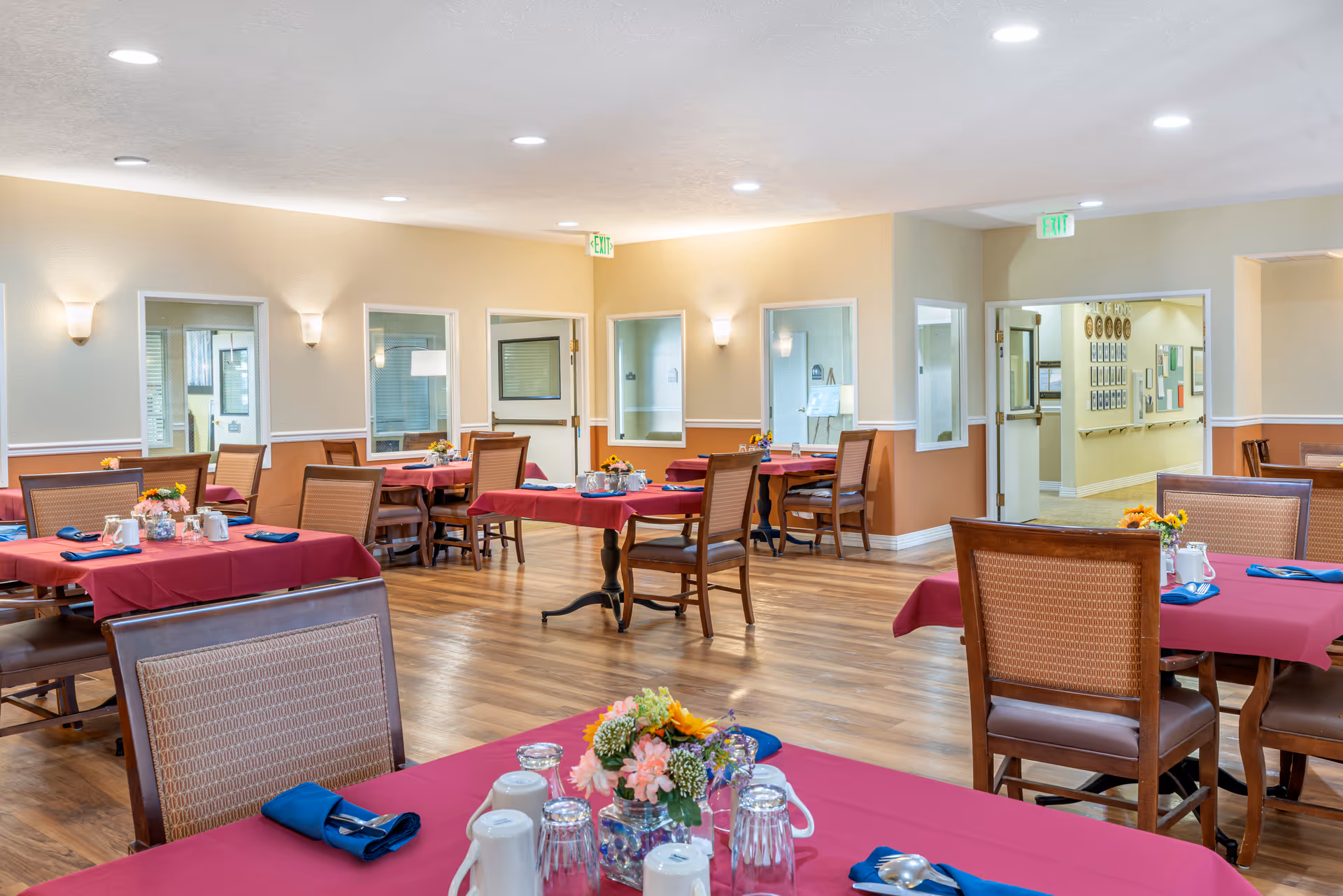 A dining room in a senior living facility with multiple tables covered in red tablecloths, each set with blue napkins, glasses, and small floral centerpieces. The room has wooden flooring, beige and light yellow walls, and several windows and doors leading to other areas.