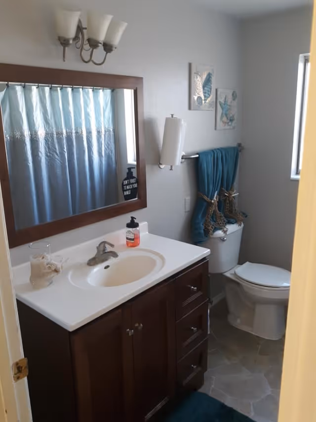 A small bathroom with a white sink and dark wood vanity, a large mirror above the sink, a toilet with teal towels hanging on a rack above it, a shower with a blue and white curtain, and a window letting in natural light.