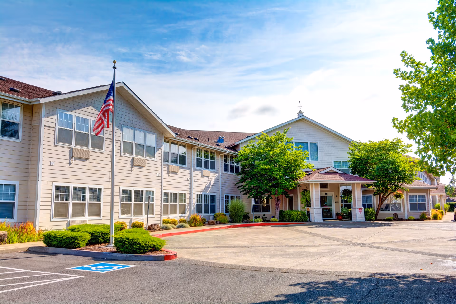 Exterior view of Cedar Village by Cogir, a two-story senior living facility with beige siding and multiple windows. There is an American flag on a flagpole in front of the building, a covered entrance with a small porch, and landscaped greenery including bushes and trees. The parking area includes a designated handicapped parking space.
