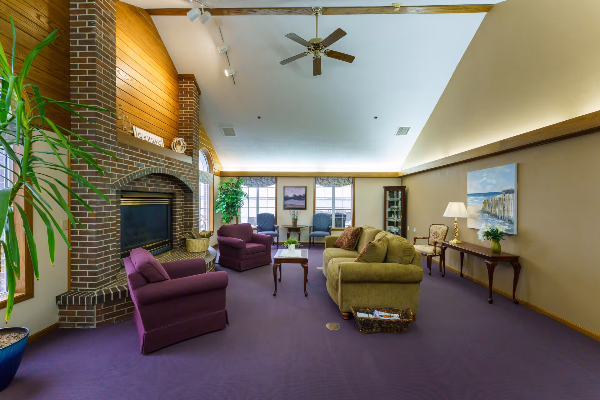 A spacious living room with a high vaulted ceiling and ceiling fan. The room features a brick fireplace with a wooden mantel, two purple armchairs, a green sofa with decorative pillows, a wooden coffee table, and a side table with a lamp and a vase of flowers. There are large windows with floral valances, a tall green plant in a blue pot, and framed artwork on the walls. The carpet is purple, and the walls are painted beige with wooden trim.