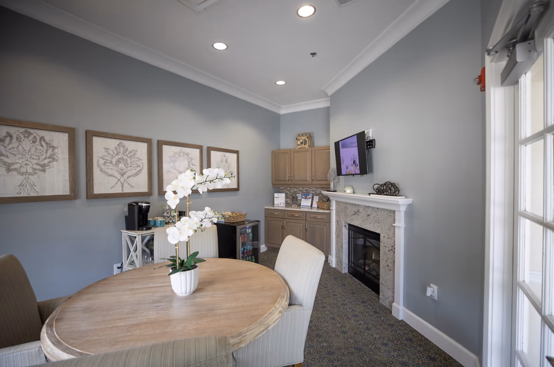 A cozy interior room with a round wooden table surrounded by upholstered chairs. A white orchid plant is placed on the table. The room has light blue walls adorned with four framed floral artworks. There is a fireplace with a mounted TV above it, and wooden cabinets with a small refrigerator and coffee maker in the corner.