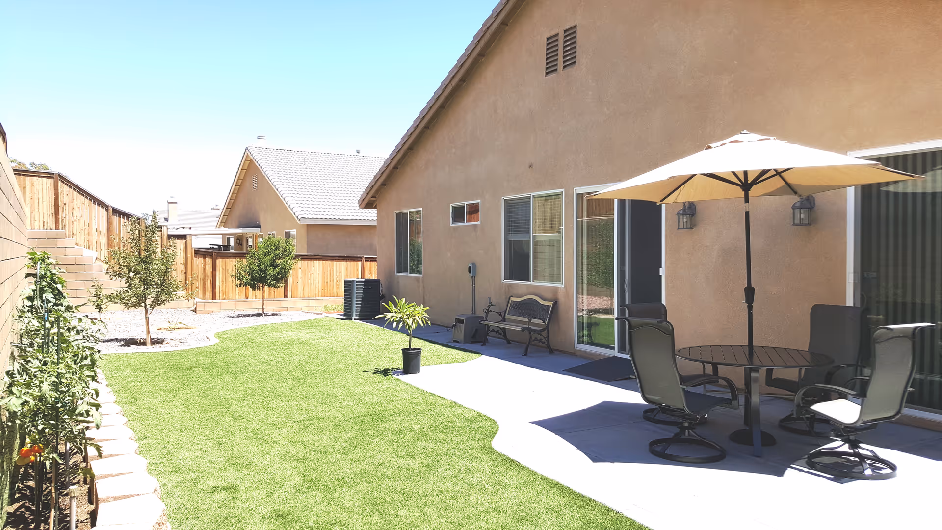 Backyard patio area of a residential facility with a beige stucco exterior wall, a round glass table with four chairs under a large beige umbrella, a bench, potted plants, small trees, and a well-maintained lawn bordered by a wooden fence and a brick wall.
