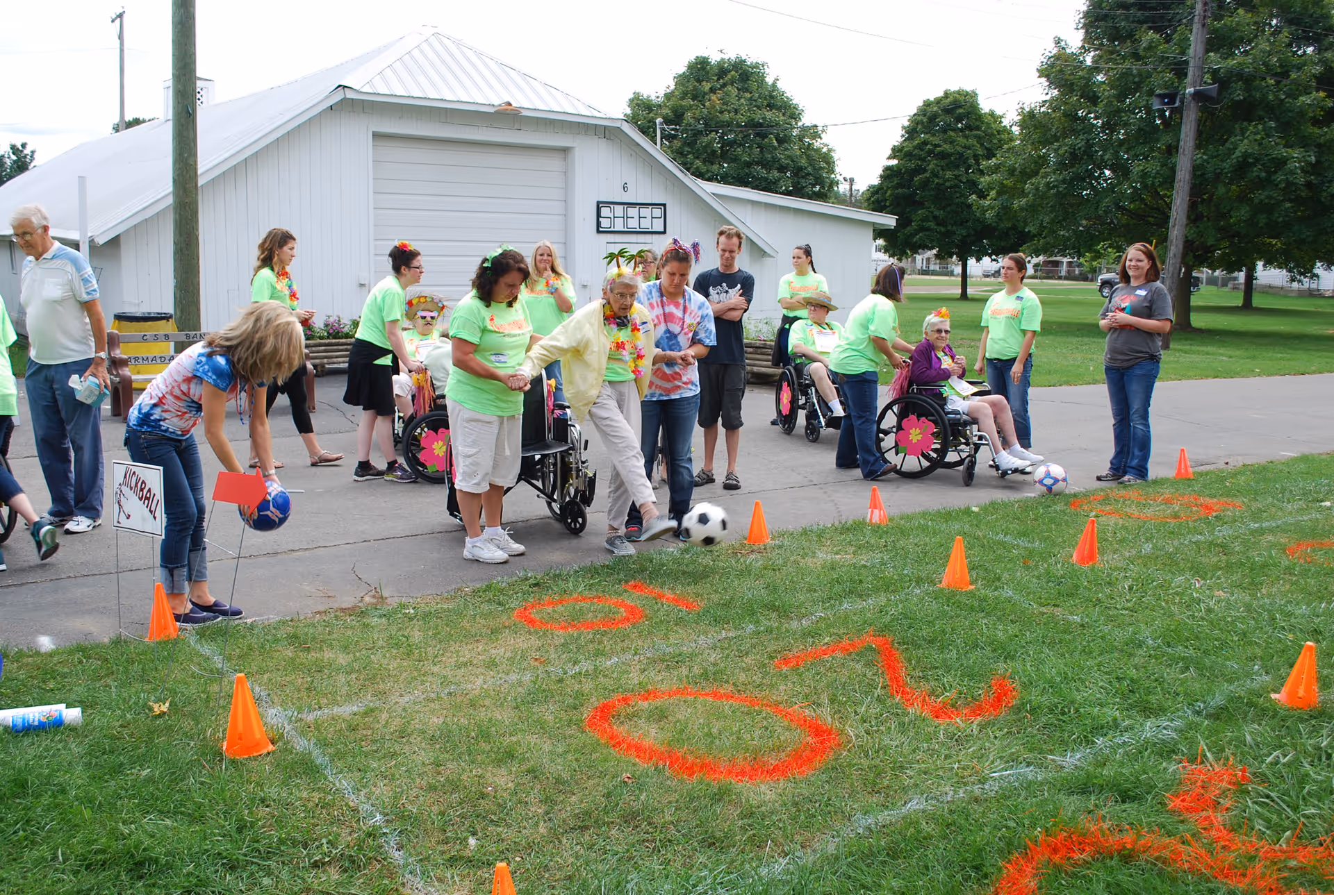 Seniors and caregivers in bright shirts play a kickball/soccer-style game outdoors with cones and spray-painted numbers in front of a white building.