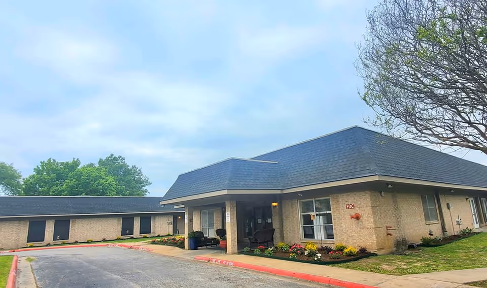 Front entrance of a one-story brick nursing facility with a covered drop-off, driveway and landscaped flowerbeds.