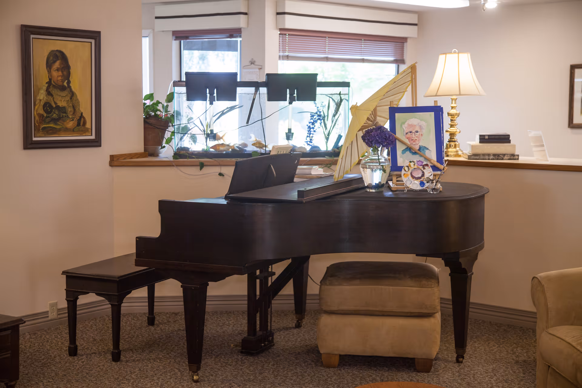 A cozy interior corner featuring a black grand piano with a matching bench and a beige ottoman. On top of the piano are decorative items including a small vase with purple flowers, a framed portrait of an elderly person, a decorative fan, and a lamp with a white shade. Behind the piano is a window with blinds and an aquarium with plants and fish. A framed painting of a young girl hangs on the wall to the left.
