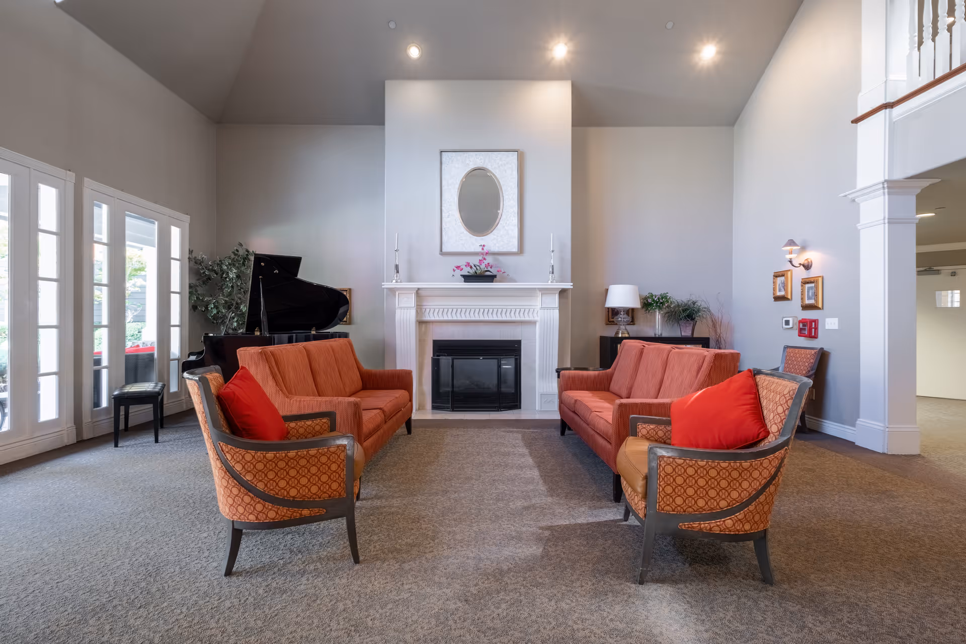 A spacious common living room with orange seating arranged around a fireplace, a grand piano, and large windows.