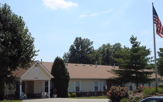 Exterior view of a single-story building with a brown roof and light-colored siding, surrounded by trees and bushes. There is an American flag on a flagpole to the right and a small covered entrance with chairs near the front door.