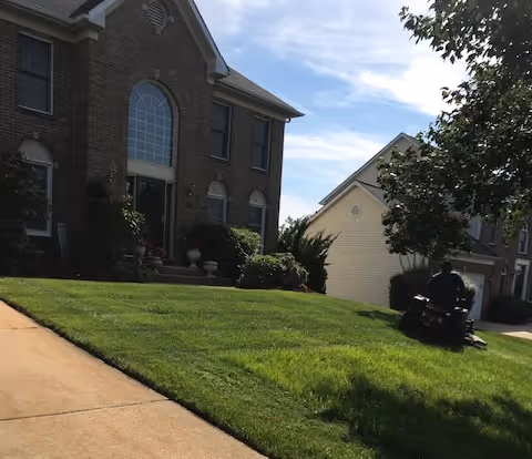 Brick senior living building with arched windows, manicured lawn, and a person near a mower.