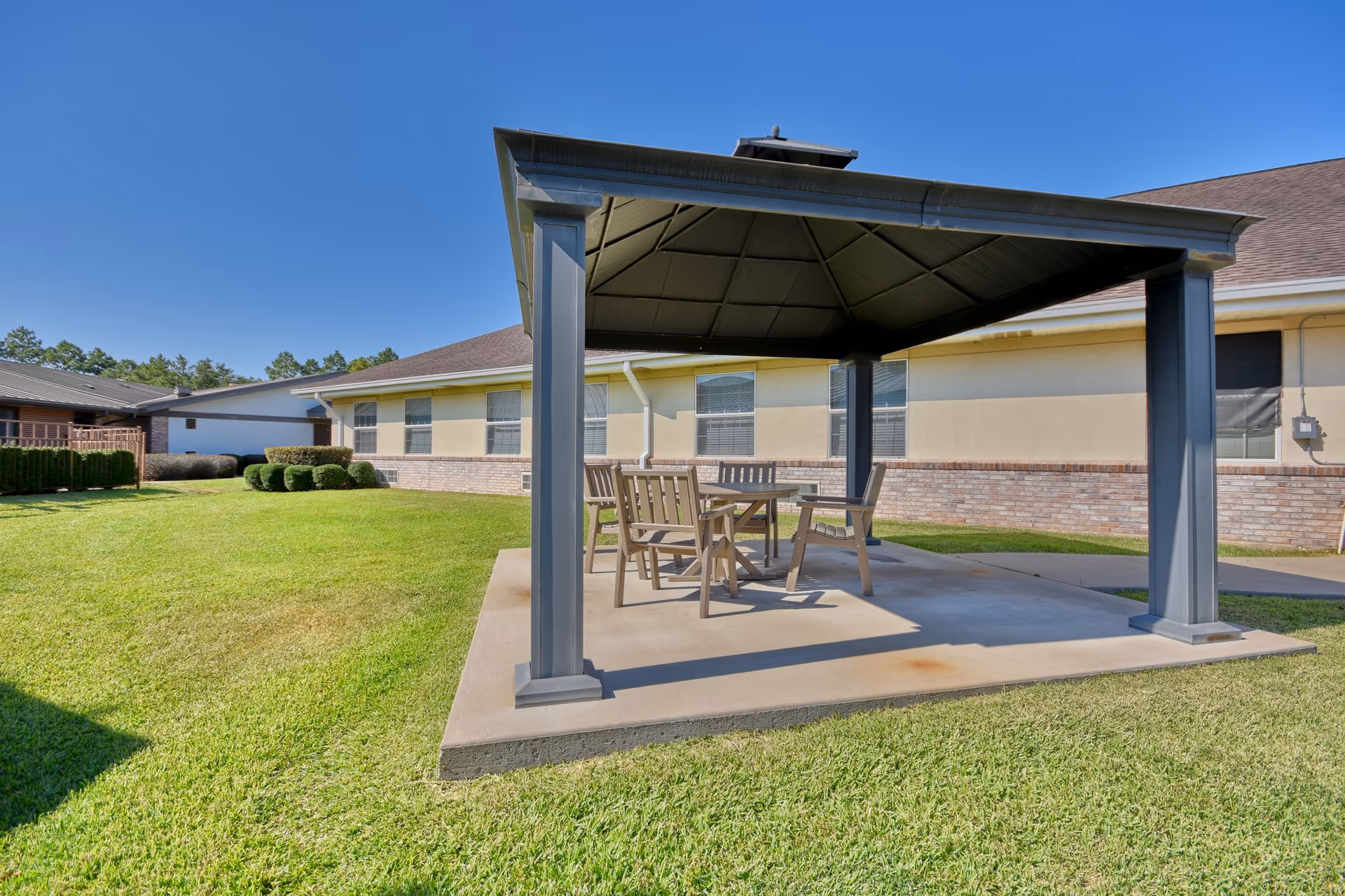 Outdoor patio area with a metal gazebo structure providing shade over a wooden table and chairs, situated on a concrete slab surrounded by green grass and bushes, with a beige building in the background under a clear blue sky.